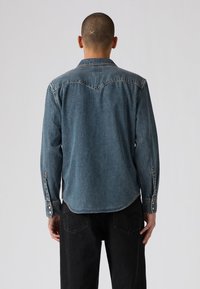 Cămașă din denim, de culoare albastru mediu, cu guler ascuțit și detalii pe spate. Mâneci lungi cu manșete cu nasturi, tiv curbat și textură uzată.