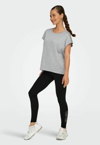 Maglietta grigia a maniche corte con una vestibilità ampia, abbinata a leggings neri con dettagli stampati e sneaker atletiche bianche.