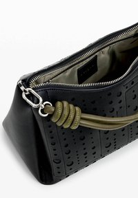 Schwarze Lederhandtasche mit durchbrochenem Design, Reißverschluss und einem olivgrünen, gewundenen Griff. Das Innere ist mit einem glatten, hellen Stofffutter ausgestattet.