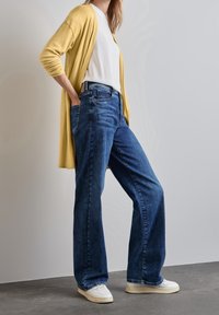 Modré džínsy s širokými nohavicemi a předními kapsami, kombinované s bílým tričkem a žlutým kardiganem, obuté s bílými teniskami. Hladký povrch po celém outfitu.