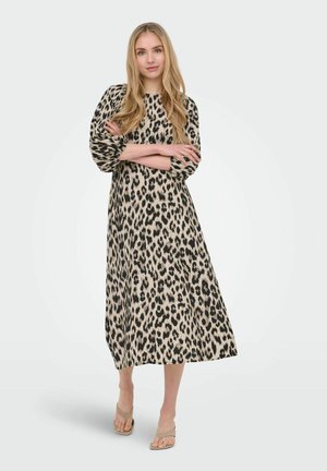 Jeune femme debout, les bras croisés, portant une longue robe beige à imprimé léopard noir et des sandales beiges sur un fond uni.