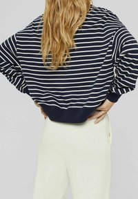 Sweat-shirt ample à rayures bleu marine avec rayures blanches et ourlet côtelé. Porté avec un pantalon clair, affichant une silhouette décontractée.