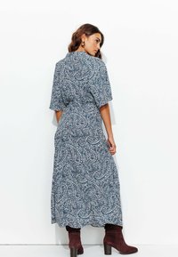 Robe maxi à motifs paisley bleus avec des manches courtes et une taille cintrée. Associée à des bottes à talons montant jusqu'aux genoux de couleur marron. Texture de tissu lisse.
