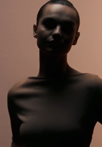 Tettpassende topp i mørk brun stoff med en glatt tekstur, med en vid utringning og korte ermer, mot en myk rosa bakgrunn.