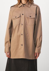 Beige knap-oe shirt med to frontlommer, klassisk krave og lange ærmer. Fremstillet af glat stof, med rundet nederste kant.