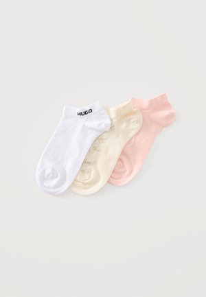 Trois socquettes blanches avec le texte "HUGO", crème avec un motif de texte subtil, et rose pastel uni, posées côte à côte sur une surface blanche.