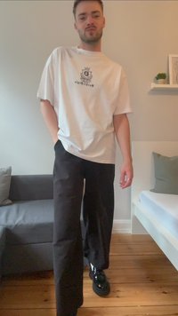 Hvit bomulls T-skjorte med logo, kombinert med løse svarte bukser og svarte sko med blank finish. Tregulv i en minimalistisk setting.