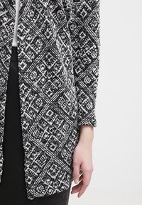 Czarny i biały kardigan w wzory z geometrycznym wzorem, wykonany z teksturowanej tkaniny, luźny krój, z otwartym przodem.