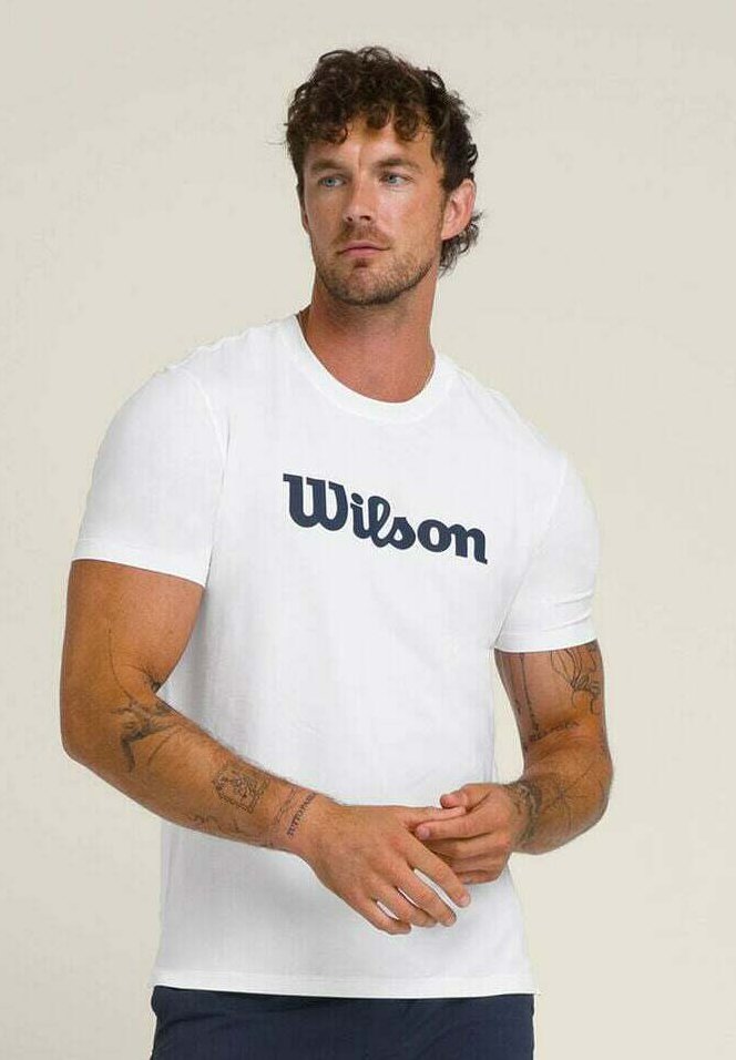 Hombre con cabello rizado, vistiendo una camiseta blanca de Wilson, con tatuajes visibles en los brazos, de pie frente a un fondo claro y sencillo, con las manos entrelazadas.