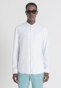 Camicia bianca con colletto a fascia e maniche lunghe, abbinata a pantaloni azzurri chiari e occhiali da sole neri. Tessuto morbido.