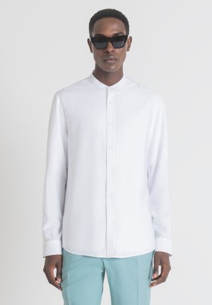 Camicia bianca con colletto a fascia e maniche lunghe, abbinata a pantaloni azzurri chiari e occhiali da sole neri. Tessuto morbido.