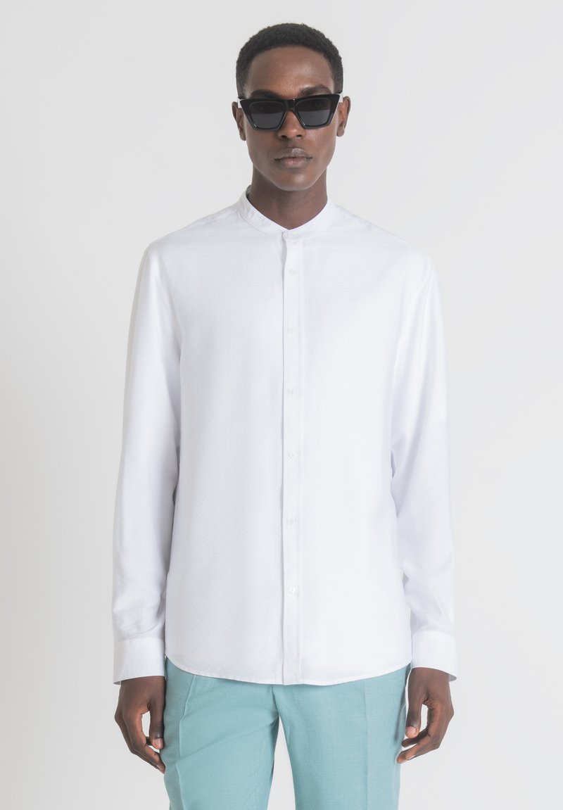 Camicia bianca con colletto a fascia e maniche lunghe, abbinata a pantaloni azzurri chiari e occhiali da sole neri. Tessuto morbido.