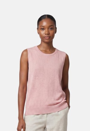 Donna con pelle scura liscia e capelli raccolti, che indossa un top rosa chiaro senza maniche e pantaloni beige chiaro, in piedi davanti a uno sfondo semplice.