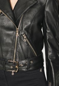 Čierna kožená motocyklová bunda s výraznými zipsami, zlatými kovovými doplnkami, širokým golierom a opaskom v páse so shining sponou.