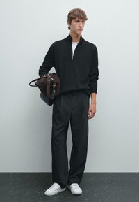 Zwarte ritstrui, witte shirt eronder, luchtige zwarte broek en witte sneakers. Houdt een bruine suède tas met leren accenten vast.