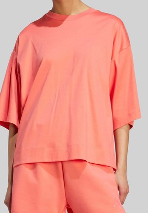 Osoba v pohodlném korálově růžovém krátkém rukávu a odpovídajících šortkách na jednobarevném šedém pozadí.