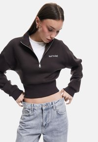 Brązowy, krótki sweatshirt z zamkiem błyskawicznym w kołnierzu i haftem „Not Today!”, zestawiony z jasnoniebieskimi, luźnymi dżinsami. Miękka tkanina, luźny krój.