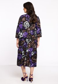 Lang blomsterkjole med mønstre af lilla, brune og hvide roser på sort baggrund. Kjolen har et lagdelt design og trekvartlange ærmer.