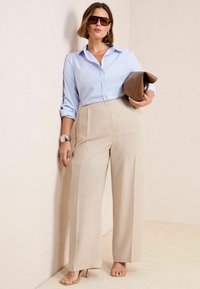 Chemise à rayures bleues et blanches, pantalon large beige, sandales beiges et sac à main marron. Le modèle porte des lunettes de soleil et une montre en argent.