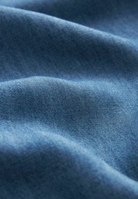 Material denim de culoare albastră medie, cu o textură uniformă, având modele țesute subtile și o cădere naturală, care evidențiază pliuri moi și flexibile.