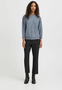 Pull gris en tricot avec motif en câble, col rond et manches trois-quarts, associé à un pantalon noir droit et des bottines.