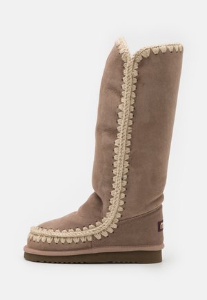 Hohe braune Wildlederstiefel mit aufwendiger beiger Stichkante, runder Zehenform und flacher Sohle. Schlichtes, geradliniges Design.