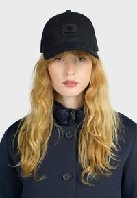Black baseball cap with a flat brim, featuring a square, embossed logo patch. The wearer has long, wavy hair, complemented by a navy blue coat.