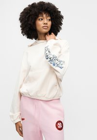 Φούτερ με κουκούλα σε κρεμ χρώμα με τυπωμένο σχέδιο στο αριστερό μανίκι, συνδυασμένο με ροζ ανοιχτό φόρμες με κεντημένες λεπτομέρειες. Χαλαρή εφαρμογή.