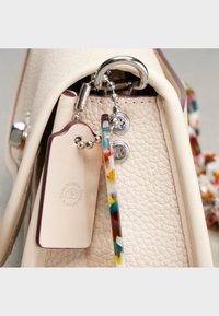 Teksturowana kremowa skórzana torebka z srebrnym pierścieniem, kolorowymi plastikowymi akcentami oraz beżową zawieszką na bagaż z logo.