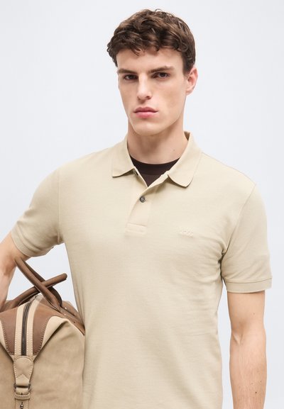 Jeune homme en polo beige portant un sac de voyage marron clair et marron foncé sur un fond pâle.