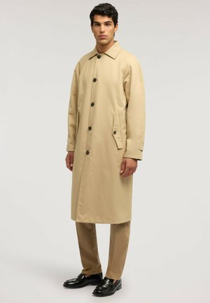 Homme portant un long manteau beige avec des boutons noirs, un pantalon beige assorti et des chaussures habillées noires, debout devant un fond clair uni.
