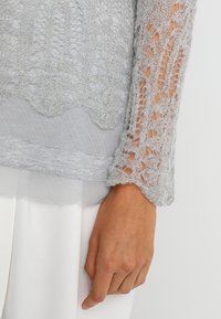 Jasnoszary dzianinowy top z misternymi koronkowymi wzorami na rękawach oraz gładkim, warstwowym wykończeniem na dole. Miękka tekstura i luźny fason.