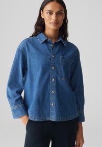 Camicia di denim con colletto, maniche lunghe, chiusura con bottoni frontali e taschino sul petto. Il tessuto è di un blu medio con una consistenza liscia.