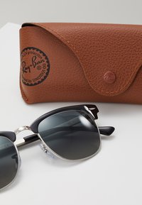 Ochelari de soare negri și argintii cu lentile degradate, având un design rotund de tip cat-eye. Însoțiți de un toc din piele maro texturată.