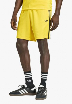Uomo che indossa pantaloncini gialli Adidas con strisce nere, calzini neri Adidas con strisce bianche e sneakers nere Adidas con strisce bianche.