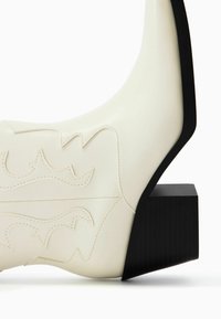 Białe skórzane botki za kostkę z zakrzywionym wzornictwem, tłoczonymi wzorami na powierzchni oraz czarnym geometrycznym obcasem w kształcie bloku.