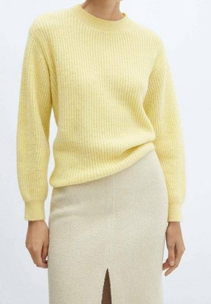 Kvinde iført en bleg gul ribstrikket sweater og et lysebeige skørt med frontslids mod en ensartet hvid baggrund.