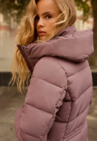 Vadderad jacka i mauve, med hög krage och sydda sektioner. Materialet ser glansigt ut, med en varm och isolerad design.