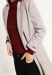 Beige wollen jas met een op maat gemaakt ontwerp, brede revers en knoopaccenten, gecombineerd met een gestructureerde rode coltrui en zwarte broeken.