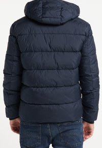 Marinblå vadderad jacka med huva, med horisontella sömmar, en slät yta och elastiska muddar. Bärs över jeans.