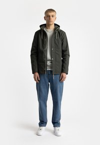 Donkergroene, waterdichte jas met een capuchon, drukknoopsluitingen en trekkoorden, gedragen over een lichtgrijze sweatshirt en blauwe jeans. Witte sneakers.