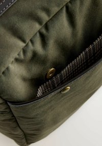 Oliivivärvi kott, millel on tekstuuriga kangas, tumeda nahkservaga ja ruudulise sise-taskuga, mis on kinnitatud metallist nupuga.