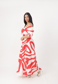 Maxikleid in Weiß mit kräftigen roten, gewellten Mustern, schulterfreies Design, gesmokter Brustbereich und leichtem, glattem Stoff. Sichtbare Absatzsandalen.