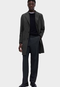 Manteau en laine gris avec un col cranté, une fermeture à boutons et une coupe ajustée, assorti à un pantalon à fines rayures foncées et des chaussures noires.