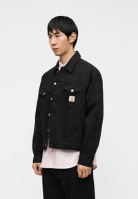 Μαύρο τζιν μπουφάν με κουμπιά μπροστά, δύο τσέπες στο στήθος και ένα μικρό λογότυπο. Ο σχεδιασμός διαθέτει άνετη εφαρμογή και κλασικό γιακά.