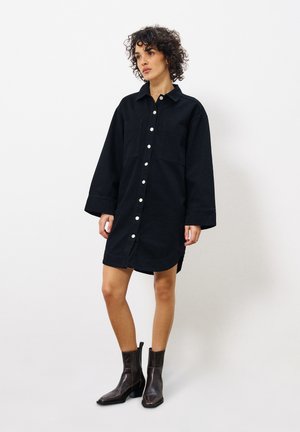 Zwart denim overhemdjurk met lange mouwen, twee borstzakken en een gebogen zoom. Model gestyled met bruine enkellaarsjes.