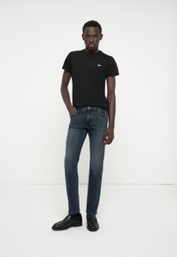 Svart t-skjorte med hvit logo, mørkeblå jeans og svarte pensko. Modellen står med hendene i lomma foran en hvit bakgrunn.