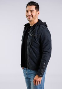 Svart läderjacka med huva, som har ett dragkedjefack och silverfärgade detaljer, bärs över en svart skjorta och blå jeans.