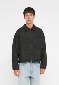 Chaqueta de rayas blancas y negras con cuello, cremallera frontal y bolsillo en el pecho; cuenta con dos bolsillos laterales y un diseño corto.