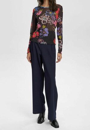 Femme portant un haut noir transparent à manches longues à motifs floraux, un pantalon large bleu marine et des bottines noires à talons, debout sur fond blanc.